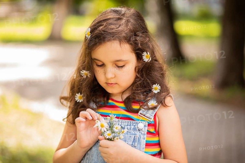 A little girl in a colorful outfit holds chamomile flowers, enjoying a sunny day outdoors. She embodies innocence surrounded by nature's beauty