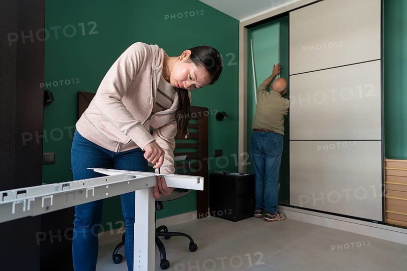 An Asian woman focuses on assembling a white desk, using tools in a modern room with green walls. In the background, a man is installing or adjusting a sliding door wardrobe panel