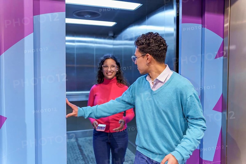 A Gen Z man holds the elevator door open for a happy woman holding a smartphone. They are casually dressed and smiling, showcasing modern social etiquette and connection