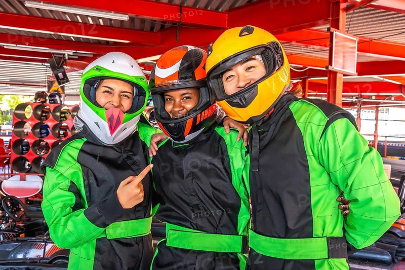 Three smiling diverse friends wearing helmets and racing suits, standing closely together at a karting track while preparing for an exciting go karting race, embracing fun and friendship