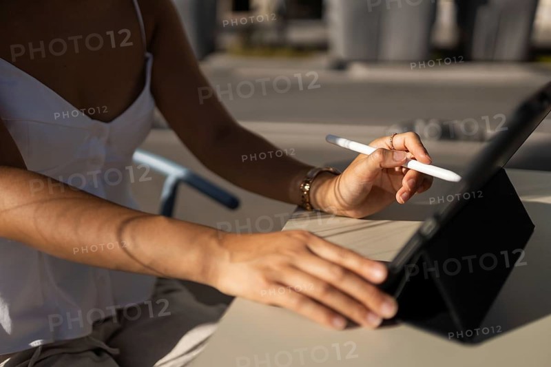 A woman in a summer outfit using a digital tablet with a stylus outdoors in a city setting. The scene captures a blend of technology and urban lifestyle