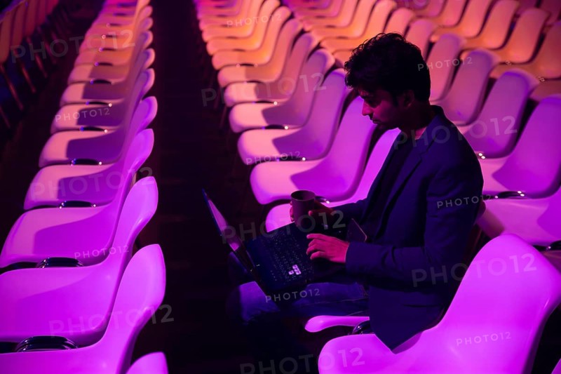 A indian man sits alone in an auditorium with purple lighting at a conference of AI in Amsterdam, working on a laptop and holding a coffee cup