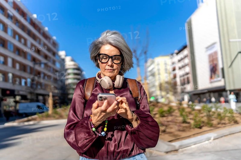 Woman with grey hair and headphones around her neck focusing on her smartphone screen while walking outdoors in a sunny urban environment, embodying modern senior travel and connection