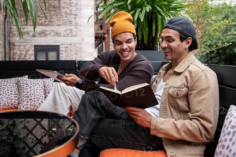 A gay couple sits on a cozy patio, sharing a joyful moment with a book. Surrounded by lush plants and soft pillows, they enjoy each other's company and literature