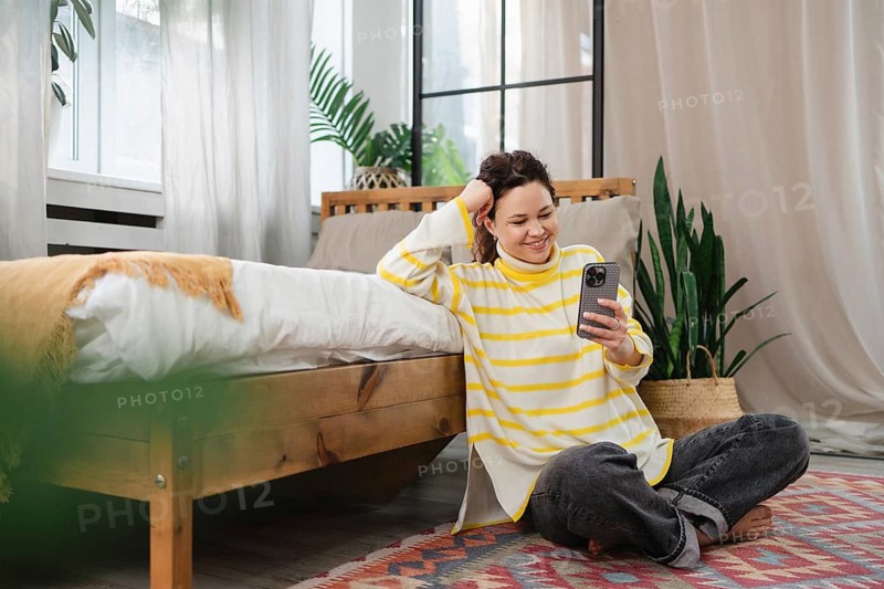 A cheerful woman, sitting on the floor in a cozy room, smiles at her smartphone. The setting includes a bed, plants, and soft light through sheer curtains, creating a relaxing vibe