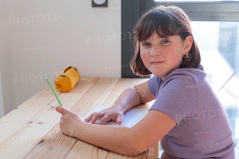 A young girl engaged in her homework sits at a wooden table, sporting a casual purple top and a focused expression