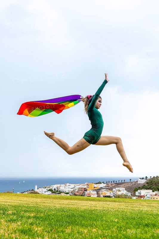 A woman dancing energetically in an open field with a rainbow flag representing LGBTQIA+ pride. The dancer's leap embodies freedom and expression against a scenic backdrop