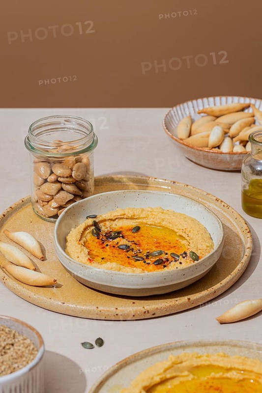 Creamy hummus garnished with seeds and spices, served on a rustic plate with breadsticks. A jar of almonds adds a crunchy touch. Perfect for Mediterranean cuisine themes