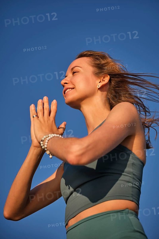 A woman practices yoga at the beach during sunset, holding hands in Namaskar Mudra while enjoying the moment