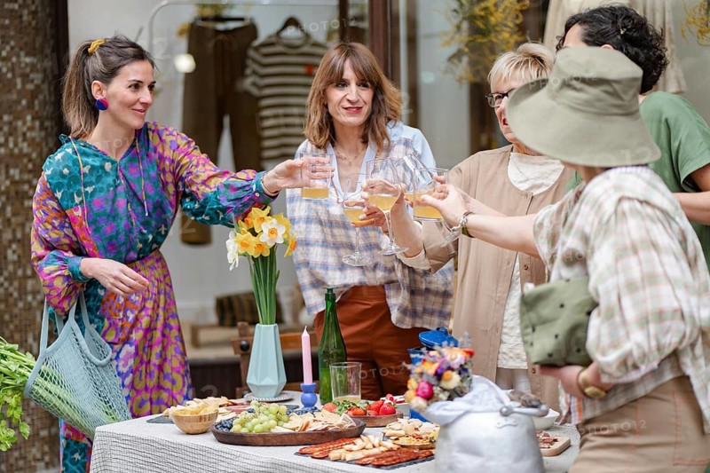 A group of coworkers enjoy an outdoor gathering, raising glasses in a celebratory toast. The table is filled with snacks, fruit, and flowers, creating a lively, inviting scene