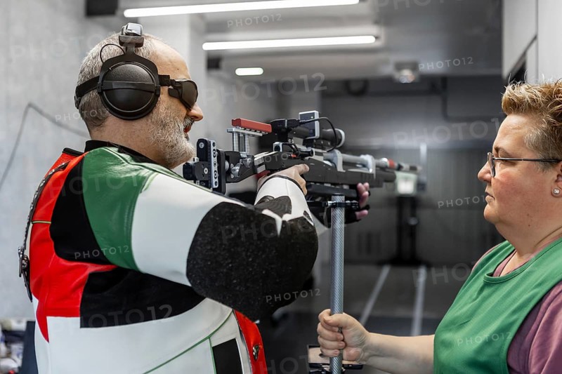 A blind athlete, wearing specialized gear, participates in a shooting competition. Guided by an assistant, showcasing skill and precision in an indoor shooting range
