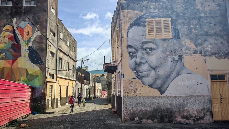 A woman with a red backpack walks through a street with a large mural of the singer 'Cesaria Evora', barefoot diva, morna, and geometric shapes, Ribeira Grande, Santo Antao, Cabo Verde, Cape Verde