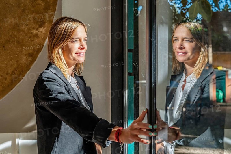 A woman stands by a window, basking in sunlight, creating a peaceful atmosphere She's dressed professionally, suggesting a remote work environment at home