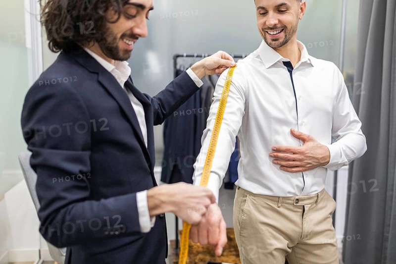 A tailor is measuring a client arm for a custom suit in a suit store. Both are smiling, emphasizing professionalism and attention to detail in men fashion and tailoring services