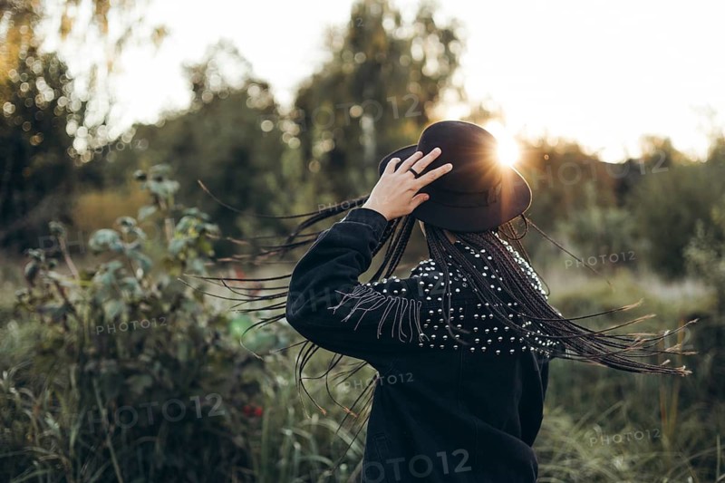 A woman stands with her back to the camera, touching her hat, as the sun sets behind her, casting a warm glow and creating a serene silhouette against a lush natural backdrop