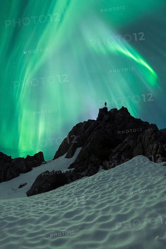 Unrecognizable person silhouette stands atop a snow-covered mountain under the vibrant green swirls of the aurora borealis on a crisp winter northern night, showcasing the awe-inspiring beauty of nature