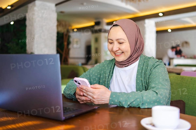 A Muslim woman wearing a hijab is smiling while using her smartphone and laptop at a cozy cafe. The atmosphere is relaxed and modern, with cultural decor