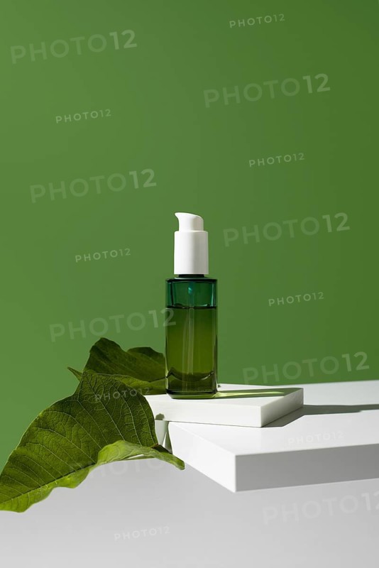 A bottle of essential oil, an ideal natural skincare product, is displayed on a white geometric stand against a solid green background, adorned with fresh leaves enhancing the theme of natural beauty