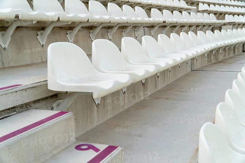 Rows of empty white seats await spectators in an outdoor athletics stadium, with visible seat numbers for organized seating