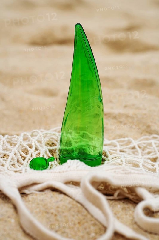 A green aloe vera gel bottle with a matching capsule lies on a beige, sandy background, intertwined with a white net bag