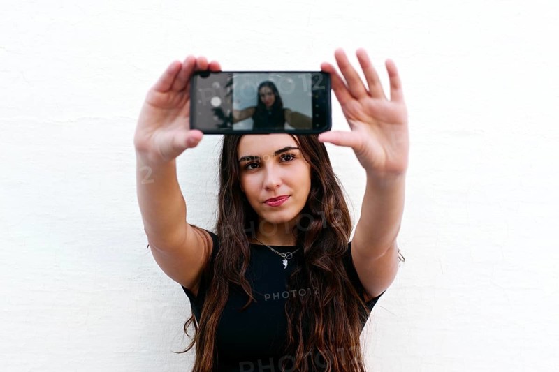 A young woman with long hair takes a selfie with her smartphone against a plain white wall. She wears a black shirt and poses casually, capturing a trendy, modern moment