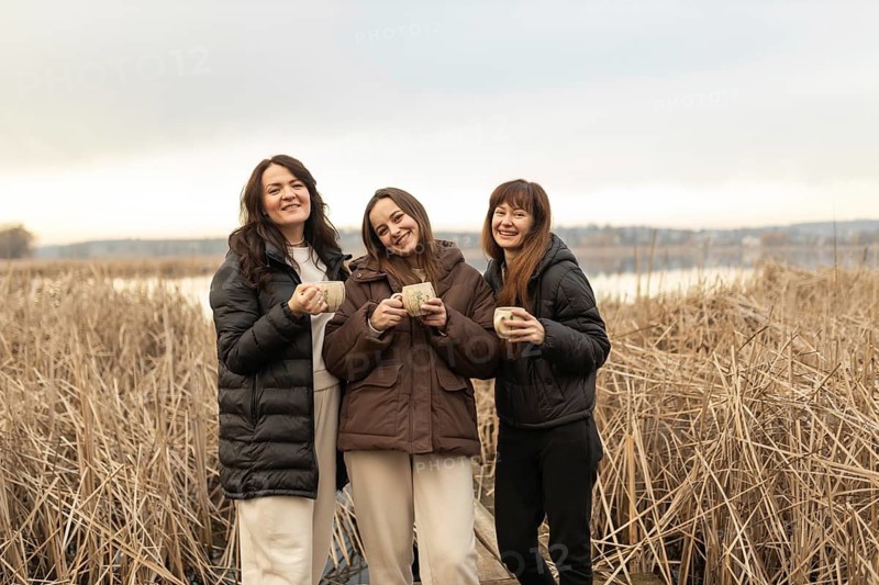 Three female friends smile joyfully as they hold mugs, standing together in a tranquil outdoor setting with tall dry reeds and a calm lake in the background, depicting warmth and camaraderie
