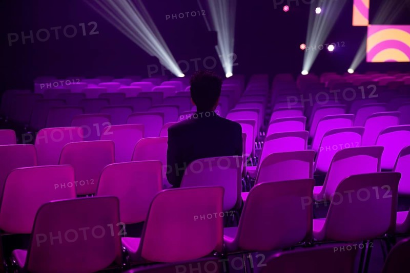 A indian man sits in an empty auditorium filled at a conference of AI in Amsterdam with purple lit chairs. The scene is illuminated by stage lights, creating a dramatic and introspective atmosphere