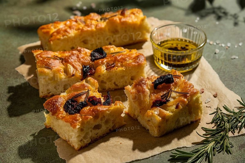 Golden homemade focaccia slices topped with olives and rosemary, accompanied by a small dish of olive oil. Perfectly presented on a rustic background, evoking Italian cuisine