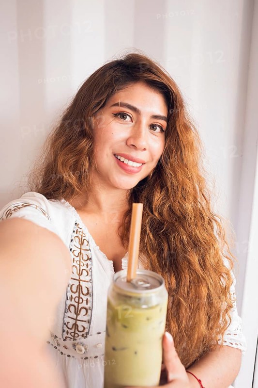 A young woman with long, wavy hair smiles while holding a refreshing iced matcha drink. She is dressed in a casual white blouse, capturing a relaxed lifestyle moment