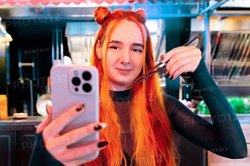 A woman with vibrant hair takes a selfie at an Asian food market, capturing her experience with chopsticks in hand The lively setting highlights cultural exploration and enjoyment