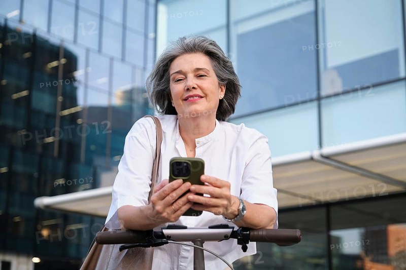 Confident mature woman in a white blouse stands outdoors, smiling while using her smartphone on an electric scooter, symbolizing connectivity and modern urban lifestyle