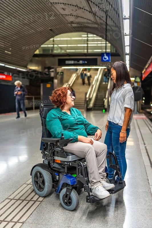 Vertical photo of a disabled woman in wheelchair talking to an asian female friend waiting at metro platform