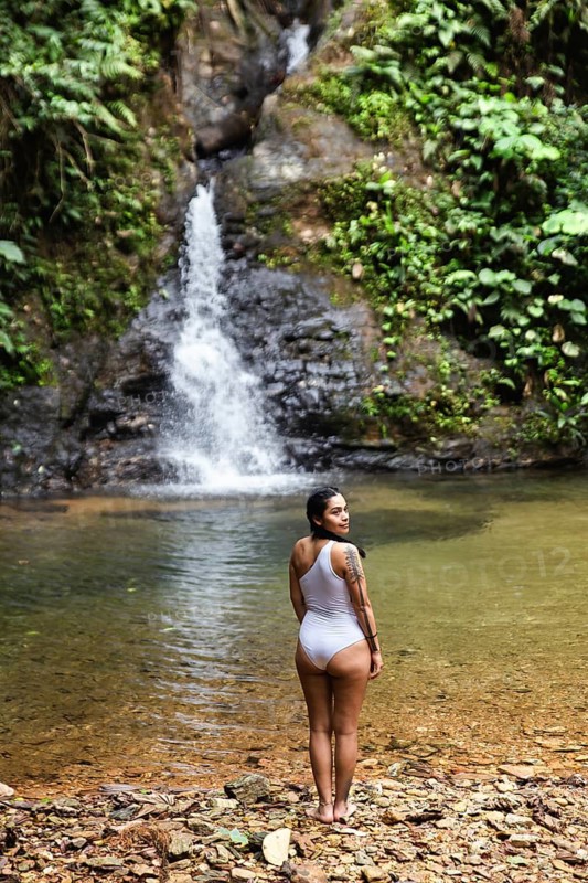 Back view of a young Ecuadorian woman in a white swimsuit stands admiring a striking waterfall in the lush Choco Andino rainforest of Ecuador