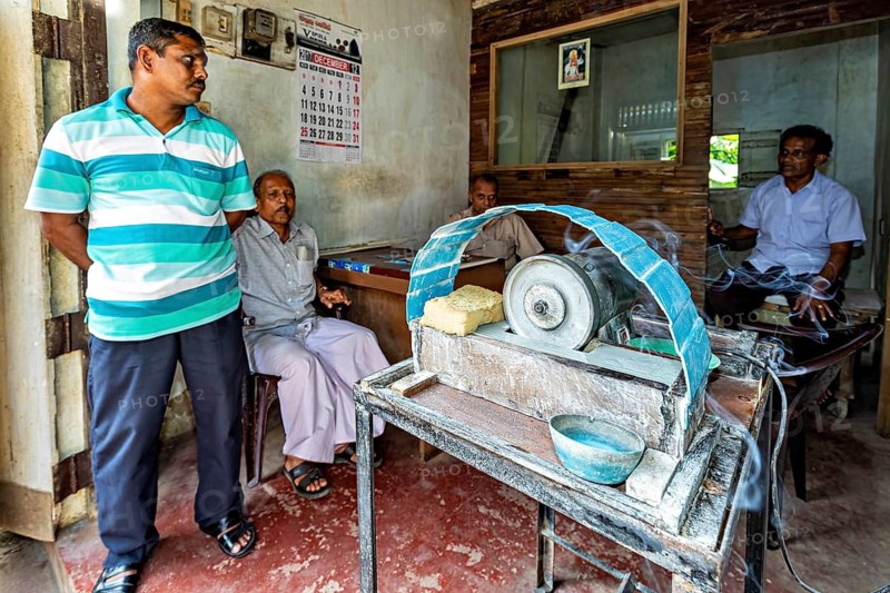 Several men are standing or sitting in a room and looking at a grinding machine, the gemstone cutters in the city of Ratnapura in Sri Lanka
