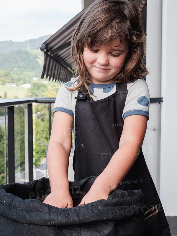 A young girl, with a gentle smile, is caught fully immersed in planting sunflower seeds in a fabric pot on a rooftop terrace. Her focused expression highlights a moment of learning and joy in urban gardening