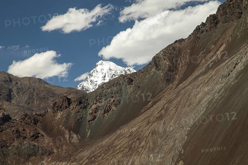 Majestic view of a snow capped mountain peak in Diskit, Nubra Valley, India, set against a backdrop of a clear blue sky with fluffy white clouds drifting by