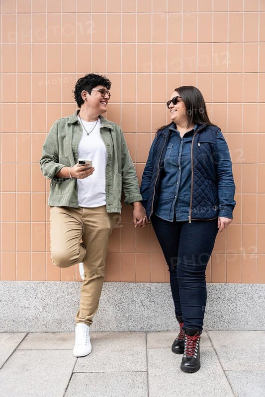 A joyful curvy couple holds hands, smiling at each other against a peach colored tiled wall. They exude happiness and connection in this urban outdoor setting