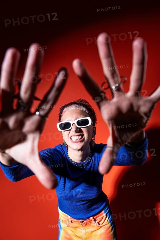 A fashionable woman poses confidently in a vivid outfit, showcasing colorful trousers, a blue top, tooth gems and trendy white sunglasses against a bold red background