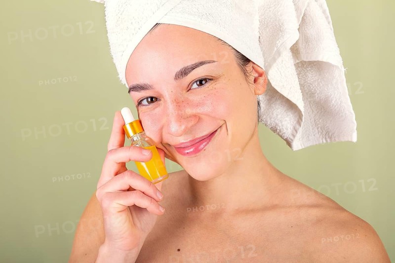 A woman with a towel wrapped around her head holds a small bottle of facial serum. Her radiant complexion and soft smile reflect a nurturing skincare routine