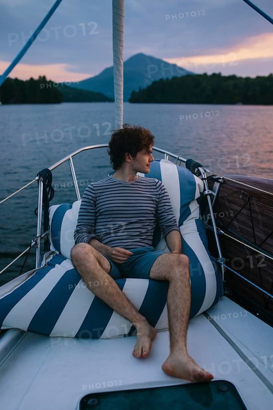 A serene afternoon on a sailboat, capturing a man relaxing against striped cushions. Surrounded by calm waters and picturesque mountains under a sunset sky