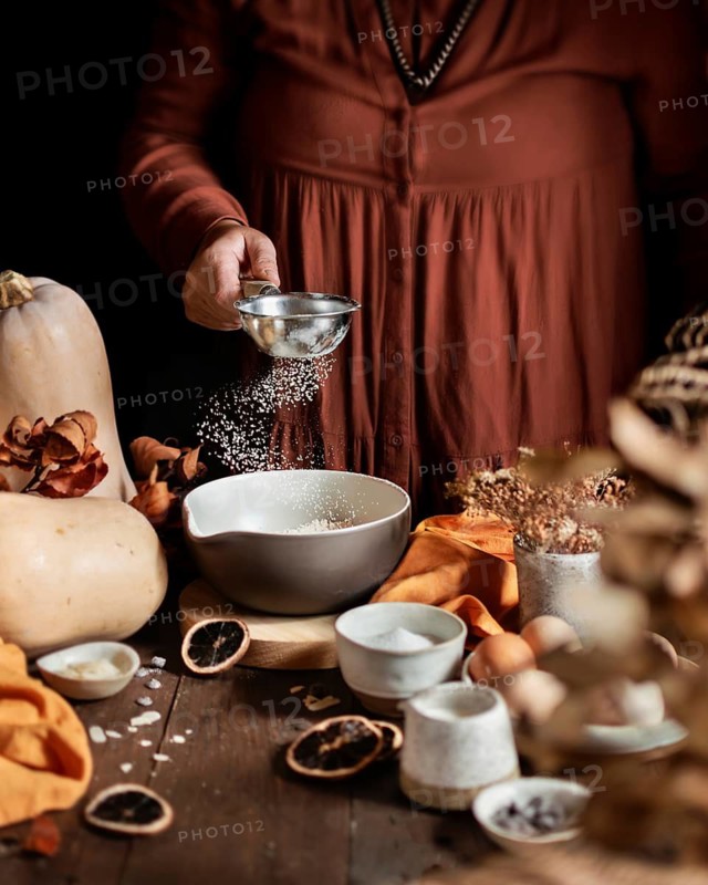 A cozy autumn scene of a homemade pumpkin cake being prepared. A person sprinkles ingredients into a bowl surrounded by pumpkins, spices, eggs, and dried fruits