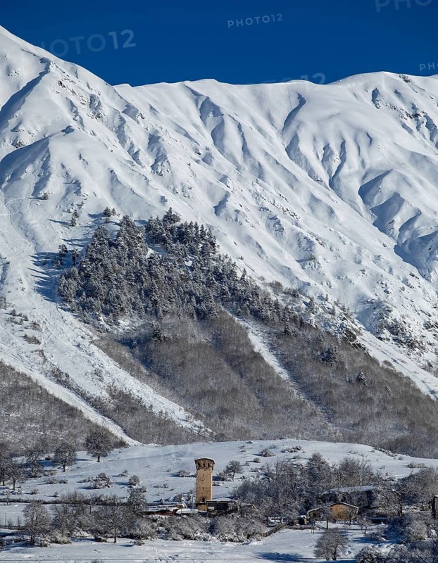 A picturesque winter scene featuring snow-covered mountains and a historic tower, surrounded by frosted trees and clear blue sky, capturing the serene beauty of a snowy landscape in Georgia