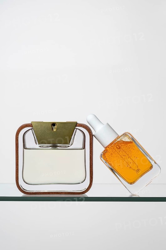 Elegant pipette bottle of vitamin C serum next to a sleek perfume bottle with a golden cap Both rest on a glass shelf, showcasing modern skincare and fragrance products