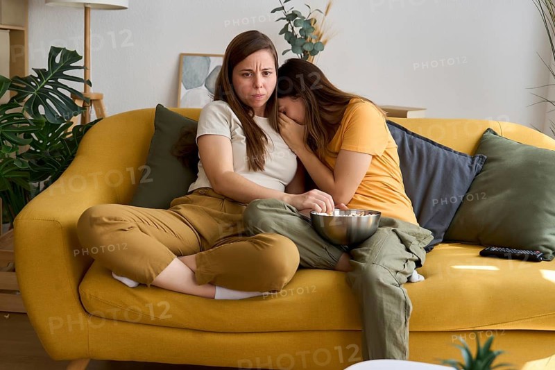 Lesbian couple on a yellow couch watching a movie One looks scared, holding some popcorn, while the other hides her face in fear The cozy living room creates a relaxed but intense atmosphere