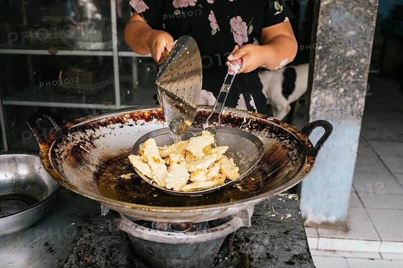 A woman fries in a large wok on a street, using a traditional strainer. The rustic setup adds an authentic touch, highlighting street food culture and culinary art