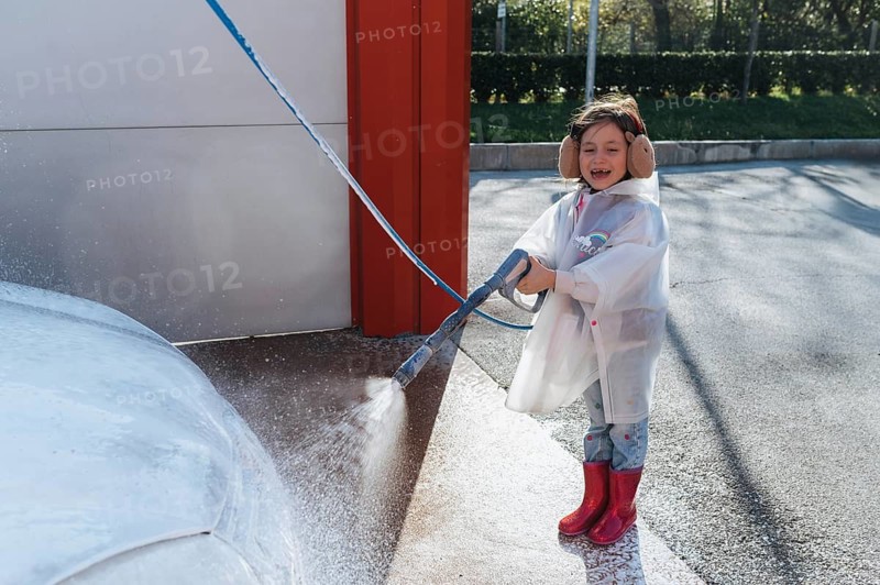 A joyful child in a raincoat and earmuffs uses a hose to spray water on a car, beaming with delight in the sunny outdoors, embracing a playful car wash adventure