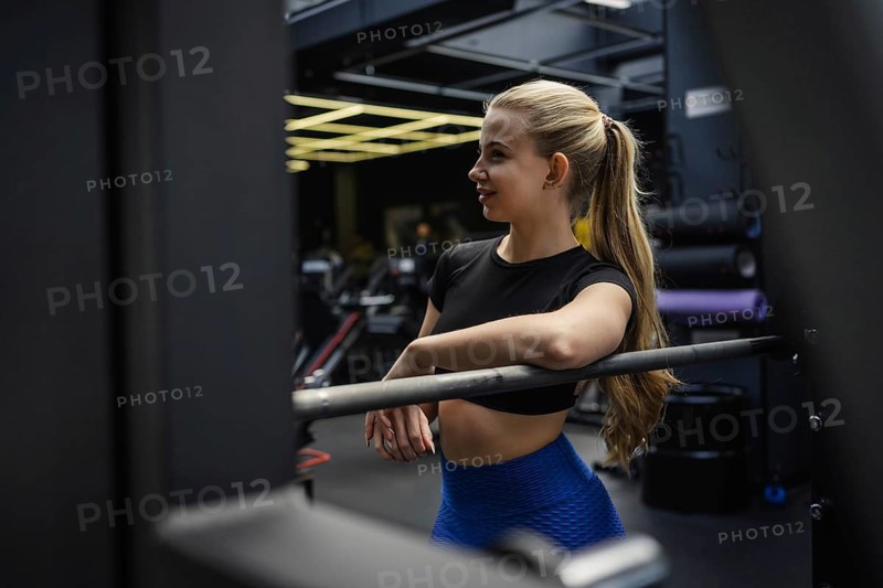 A fit blonde woman rests on gym equipment, wearing blue leggings and a black top. The gym setting includes exercise machines and a motivational ambiance
