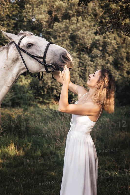 A young woman in a flowing white dress gently feeds a white horse in a sunlit forest setting, embodying harmony with nature, serenity, and intimate animal bonding