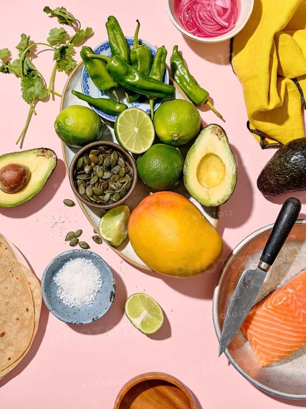 Top view of a vibrant array of taco ingredients spread on a pink backdrop, including green chilies, avocado, lime, coriander, mango, pink pickled onions, and salmon