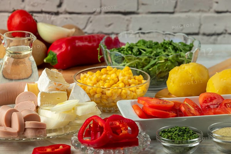 Preparation for salad with potatoes, tomatoes, peppers, sausage, corn, onions, cheese and arugula in a bright environment